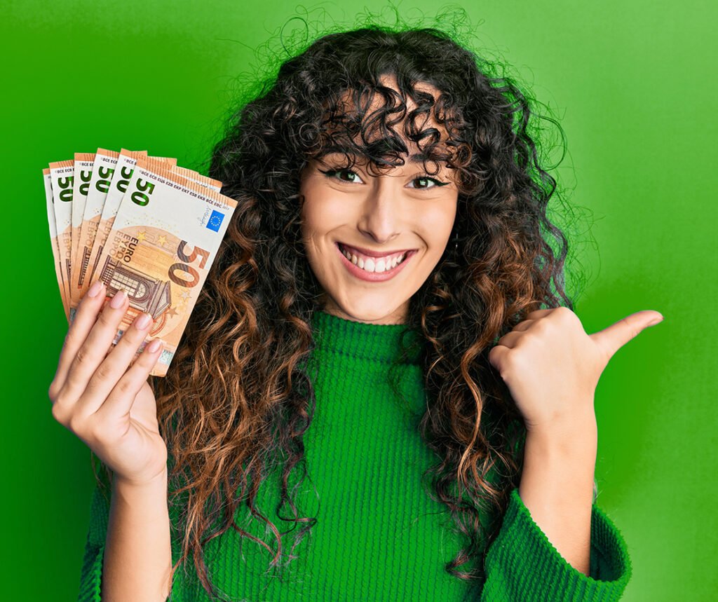 Young hispanic girl holding bunch of 50 euro banknotes pointing thumb up to the side smiling happy with open mouth
